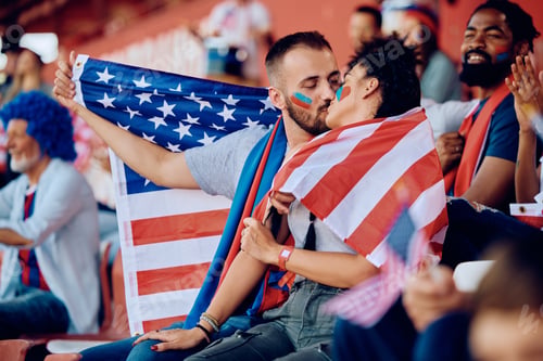 Preview: Young couple wrapped in American flag kissing during sports championship at the stadium.