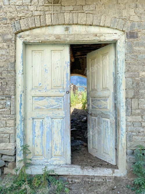 Preview: Aged wooden double door in a rustic stone wall, partially open to reveal lush greenery