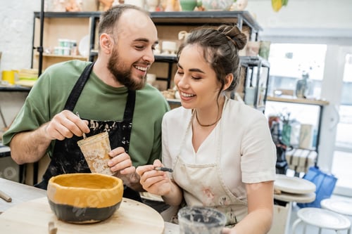 Preview: Cheerful couple of artisans holding paintbrushes whle coloring clay bowl on wooden board in studio