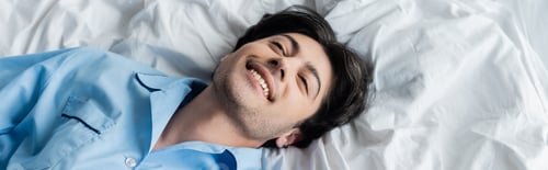 Preview: top view of young brunette man smiling while lying on white bedding, banner