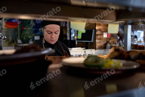 Preview: Senior chef wearing jacket arranging plates with poultry garnishes on kitchen pass-through shelf