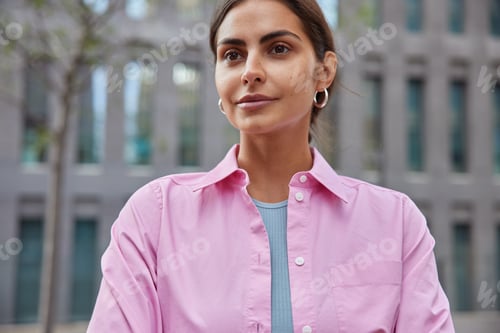 Preview: Beautiful satisfied woman with healthy skin concentrated into distance strolls in city wears pink sh