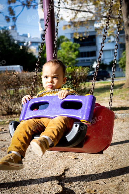 Preview: Cute baby boy having fun on outdoor playground. Toddler activities. Serious baby swinging on swing.