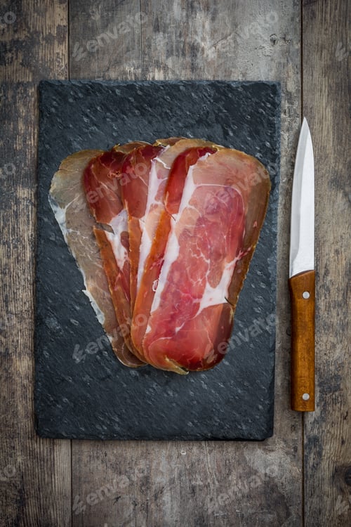 Preview: Slices of prosciutto crudo on slate cutting board with knife on rustic wooden table