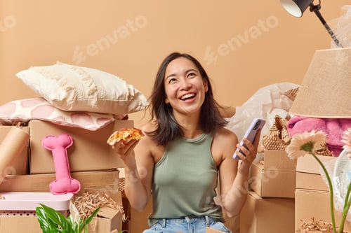 Preview: Positive young Asian woman with dark hair eats pizza holds smartphone focused above gladfully dresse