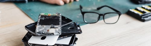 Preview: Close up view of pile of broken mobile phones near eyeglasses on workplace with blurred repairman on