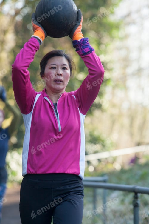 Preview: Woman in a pink jacket lifting a medicine ball overhead during an outdoor workout session.