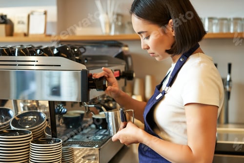 Preview: Smiling young asian barista girl, using coffee machine steamer, steaming milk for cappuccino order