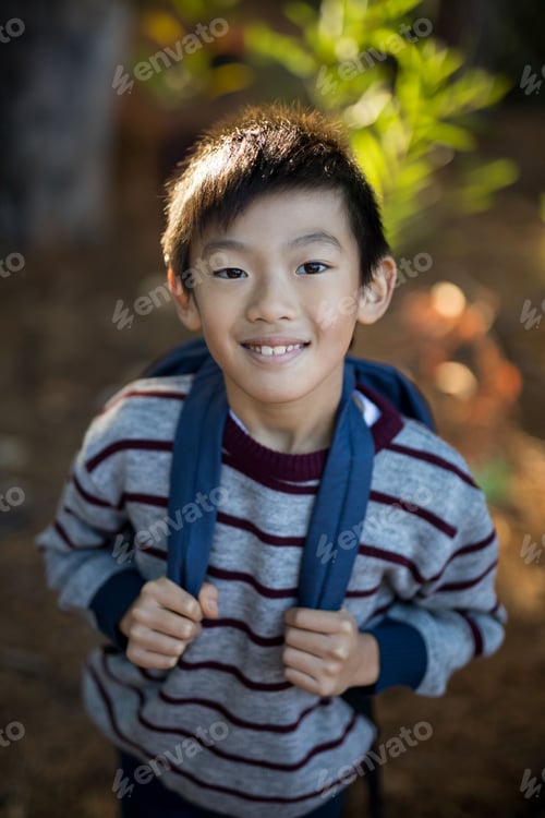 Preview: Asian boy smiling and standing on dirt path in wooded park, holding blue-strap backpack straps