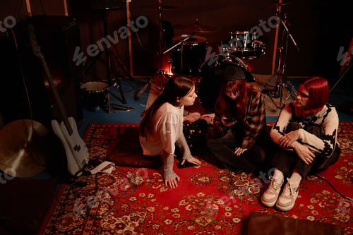 Preview: Girls Band of Three Young Artists Using Phone while Sitting on Carpet in Dim Studio