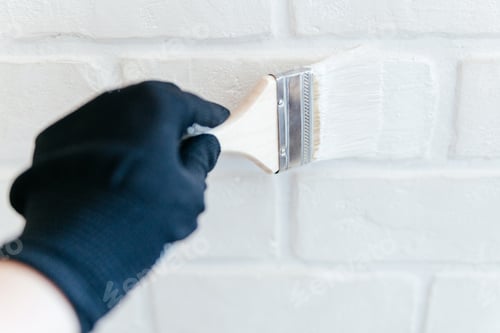 Preview: Close-up of a worker's hand with a brush painting a brick wall with white paint.
