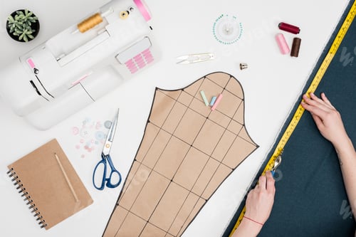 Preview: cropped shot of tailor measuring textile with measuring tape at workplace with tailoring equipment