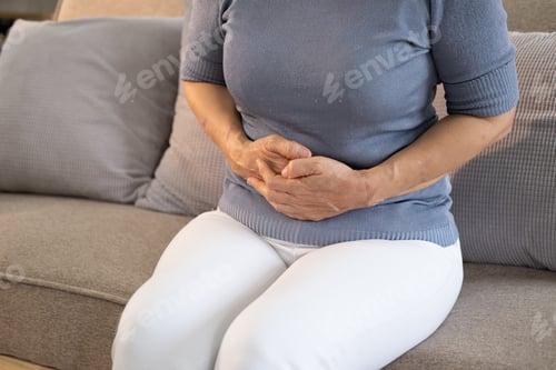 Preview: Unhealthy senior woman sit on sofa at home touch hold belly suffer from stomach ache.