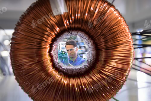 Preview: Worker inspecting electromagnetic coil seen through large coil in electromagnetics factory