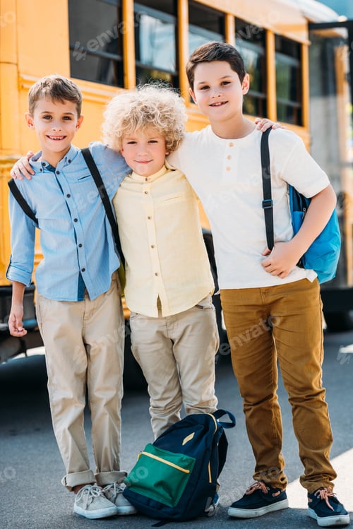 Preview: happy little schoolboys embracing in front of school bus and looking at camera