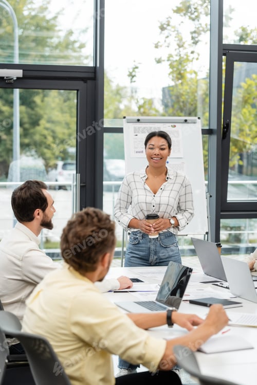 Preview: smiling multiracial woman with coffee to go talking to colleagues sitting at conference table