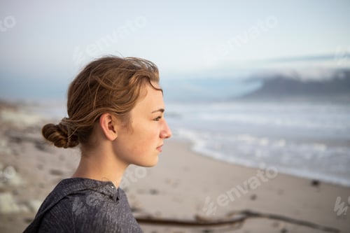 Preview: Profile portrait of a teenage girl looking out to sea from a beach at sunset.