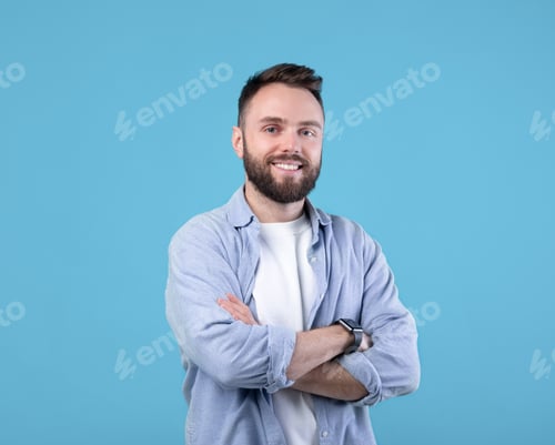 Preview: Portrait of positive young guy posing with crossed arms and smiling at camera on blue studio