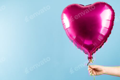 Preview: Hand holding shiny pink heart shaped balloon floating on blue background, with copy space