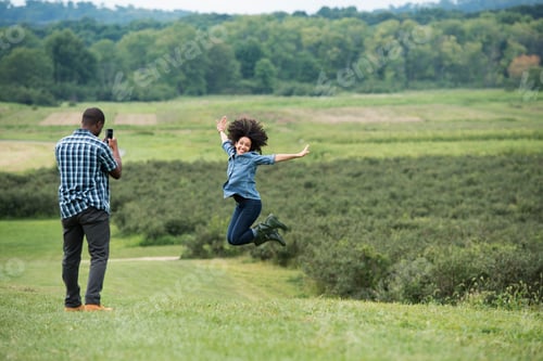 Preview: A man taking a photograph of a woman leaping in the air, jumping for joy with her arms outstretched.
