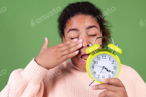 Preview: Beautiful african american woman on green background with alarm clock sleepy tired exhausted yawning