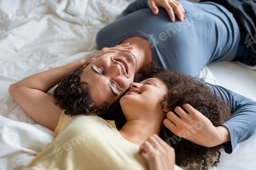 Preview: Latino young couple lying down together on bed in cozy bedroom at home.