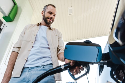 Preview: low angle view of happy man holding fuel pump while refueling automobile with benzine at gas station