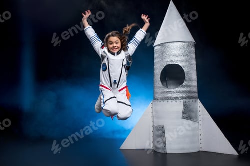Preview: Happy little girl in astronaut costume jumping near rocket