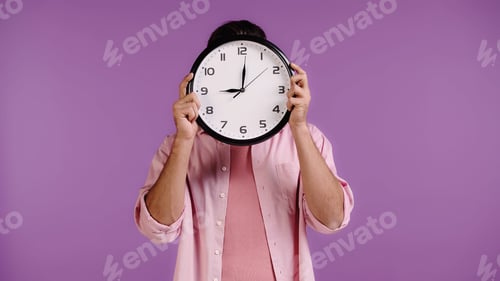 Preview: young man obscuring face with clock isolated on purple