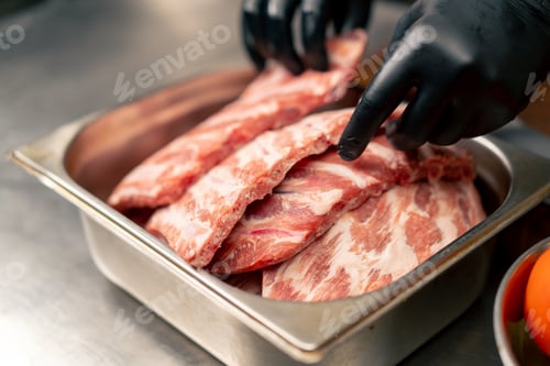 Preview: in professional kitchen hands in black gloves take ribs from a bowl and transfer them to a board