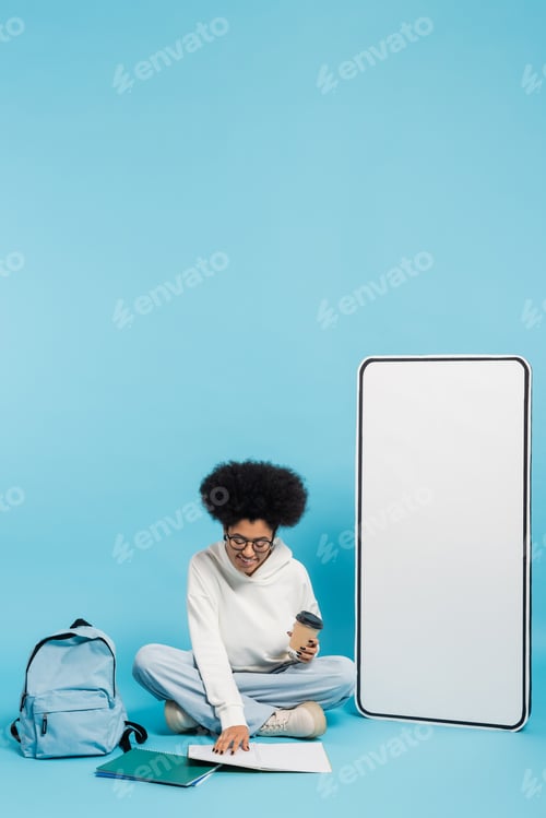 Preview: african american student with paper cup looking in notebook while sitting near backpack and white