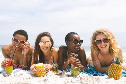 Preview: front view of multi ethnic women lying at beach while having pineapple juice on a sunny day