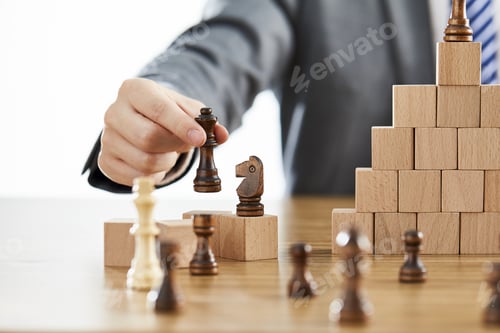 Preview: Businessman in a suit arranging chess pieces on wooden blocks on different levels