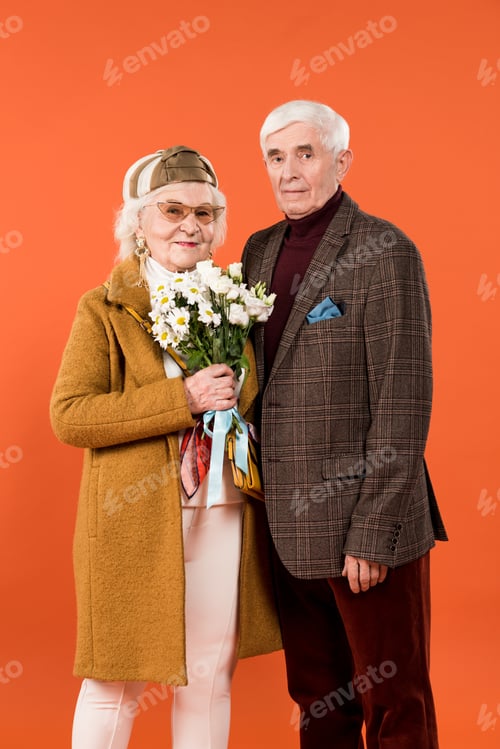 Preview: cheerful senior man standing with trendy wife holding flowers isolated on orange