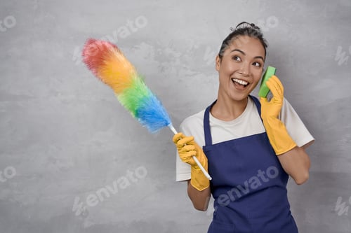 Preview: Clean with fun. Playful young woman in uniform holding colorful microfiber duster and kitchen sponge