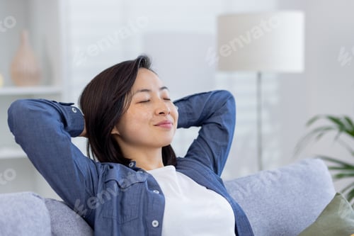 Preview: Young beautiful Asian woman relaxing at home close-up on sofa in living room, hands behind head