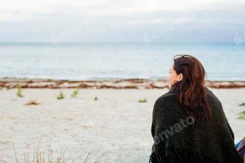 Preview: Rear view of woman wrapped in blanket sitting on beach