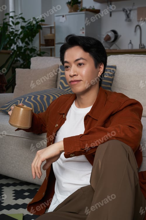 Preview: Relaxing at home in a modern apartment, an Asian man enjoys a quiet moment with tea