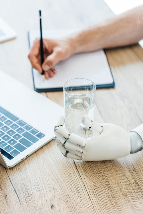 Preview: close-up view of robotic arm holding glass of water and person taking notes at wooden table