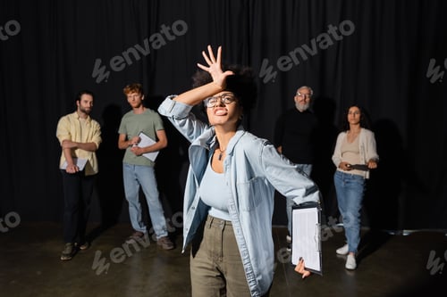 Preview: emotional african american woman grimacing and gesturing near art director and group of multiethnic