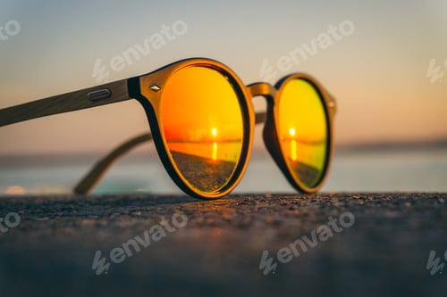 Preview: Pair of sunglasses on the beach with the reflection of the sunset