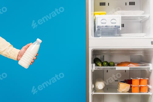 Preview: cropped view of man holding milk near open fridge and freezer with fresh food on shelves isolated on