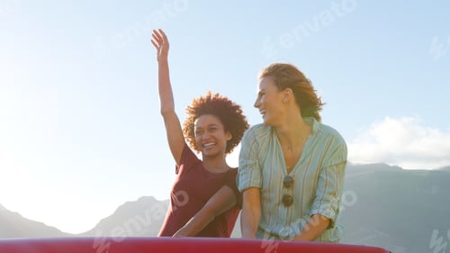 Preview: Two Female Friends Standing Up In Back Of Open Top Hire Car On Summer Vacation