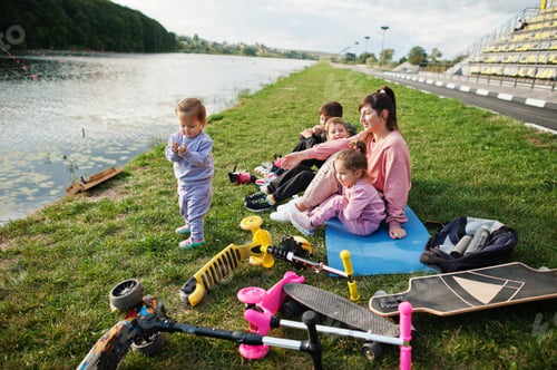 Preview: Young stylish mother with four kids sitting on grass against lake.