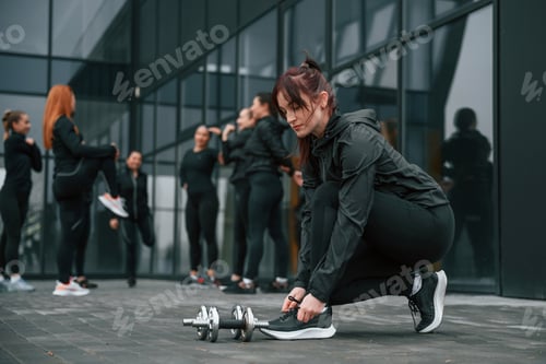 Preview: Ties shoelaces. Group of sportive women is outdoors near black building