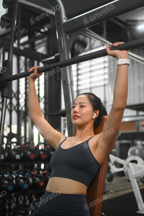 Preview: Strong sportswoman with earphones training on a smith machine in gym.