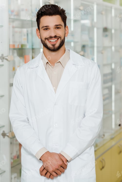 Preview: Handsome pharmacist smiling at camera by drugstore showcase