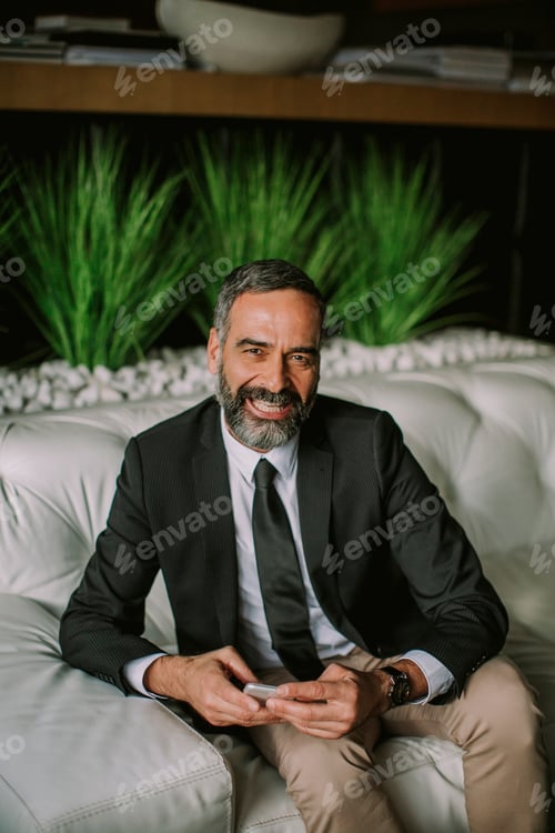 Preview: Senior businessman using mobile phone and sitting on the sofa in the office