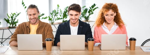 Preview: panoramic shot of three smiling friends using laptops in office