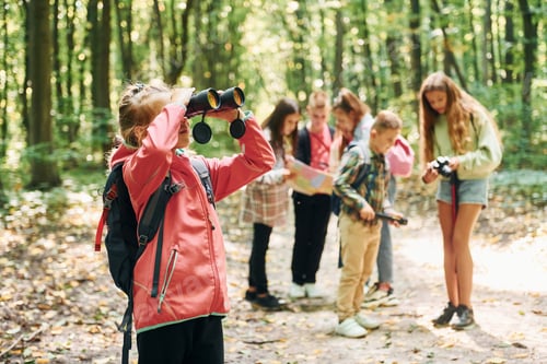 Preview: Girl looking into binoculars. Kids in green forest at summer daytime together